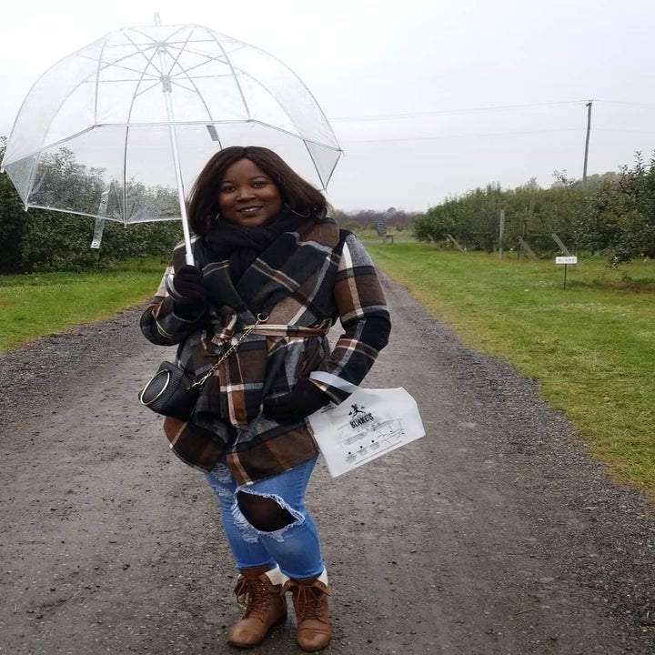 Reviewer in the brown and blue plaid coat, tied at waist, holding clear umbrella as they stand outside in middle of an orchard