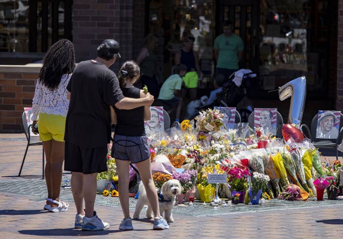 One person puts their arm around another&#x27;s shoulders as they stand near a collection of flowers and portraits