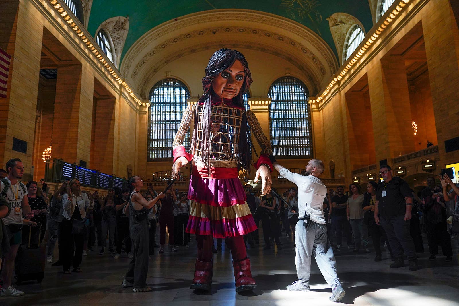 People surround a puppet of a girl, dressed in a skirt and boots and standing twice as tall as the puppeteers controlling her