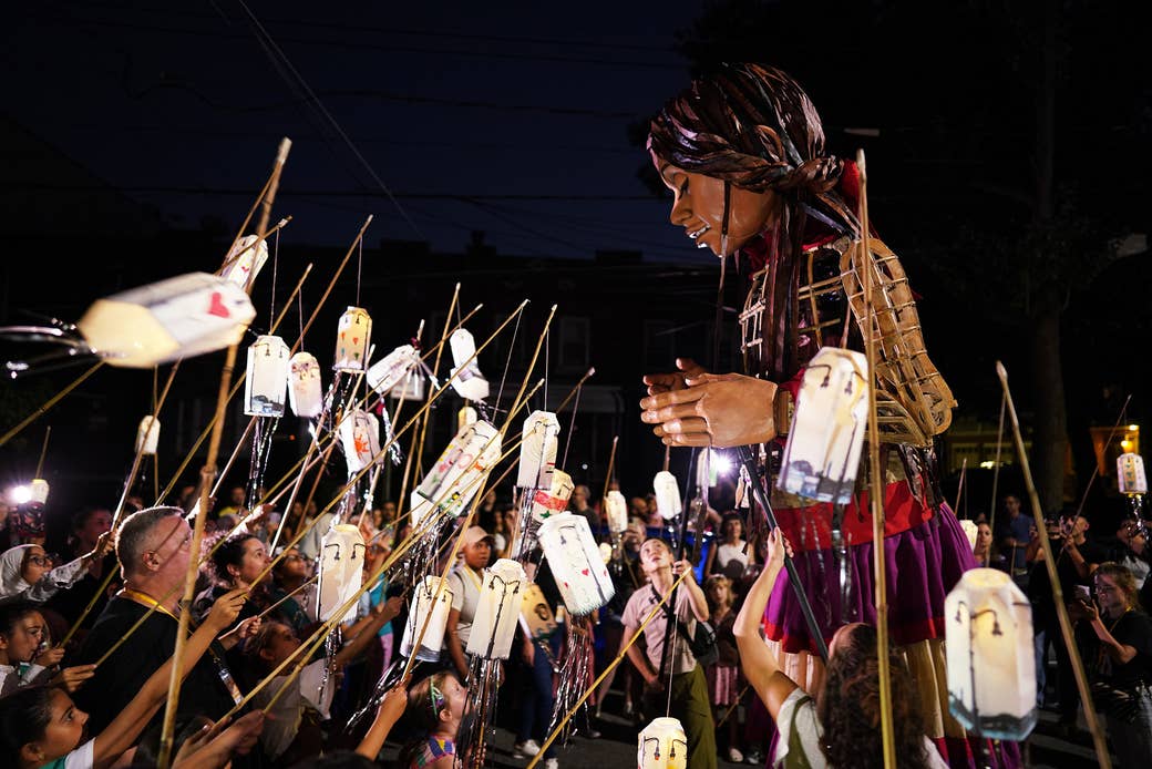 People hold up paper lanterns on sticks in front of Little Amal