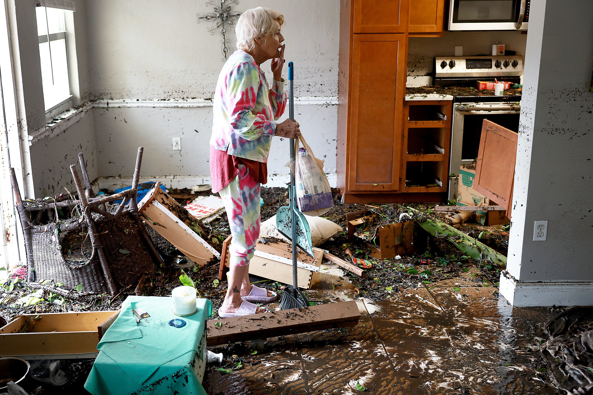 A woman holding a broom and dustpan stands in a room filled with mud and debris