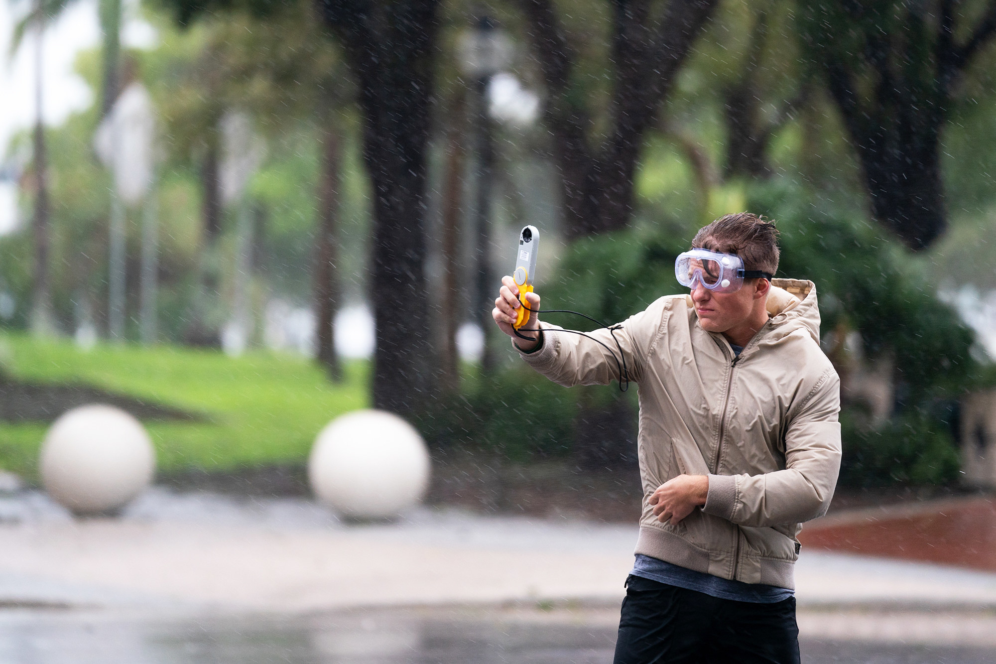 A person wearing safety goggles braces as they hold up a measurement instrument in the wind and rain