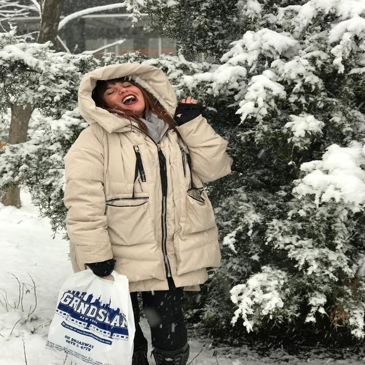 Reviewer standing in snow laughing wearing the beige coat with hood over head