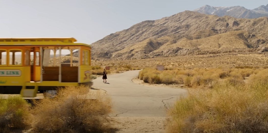 A yellow bus with &quot;Victory Town Link&quot; on the side driving out of frame as Alice runs into the desert
