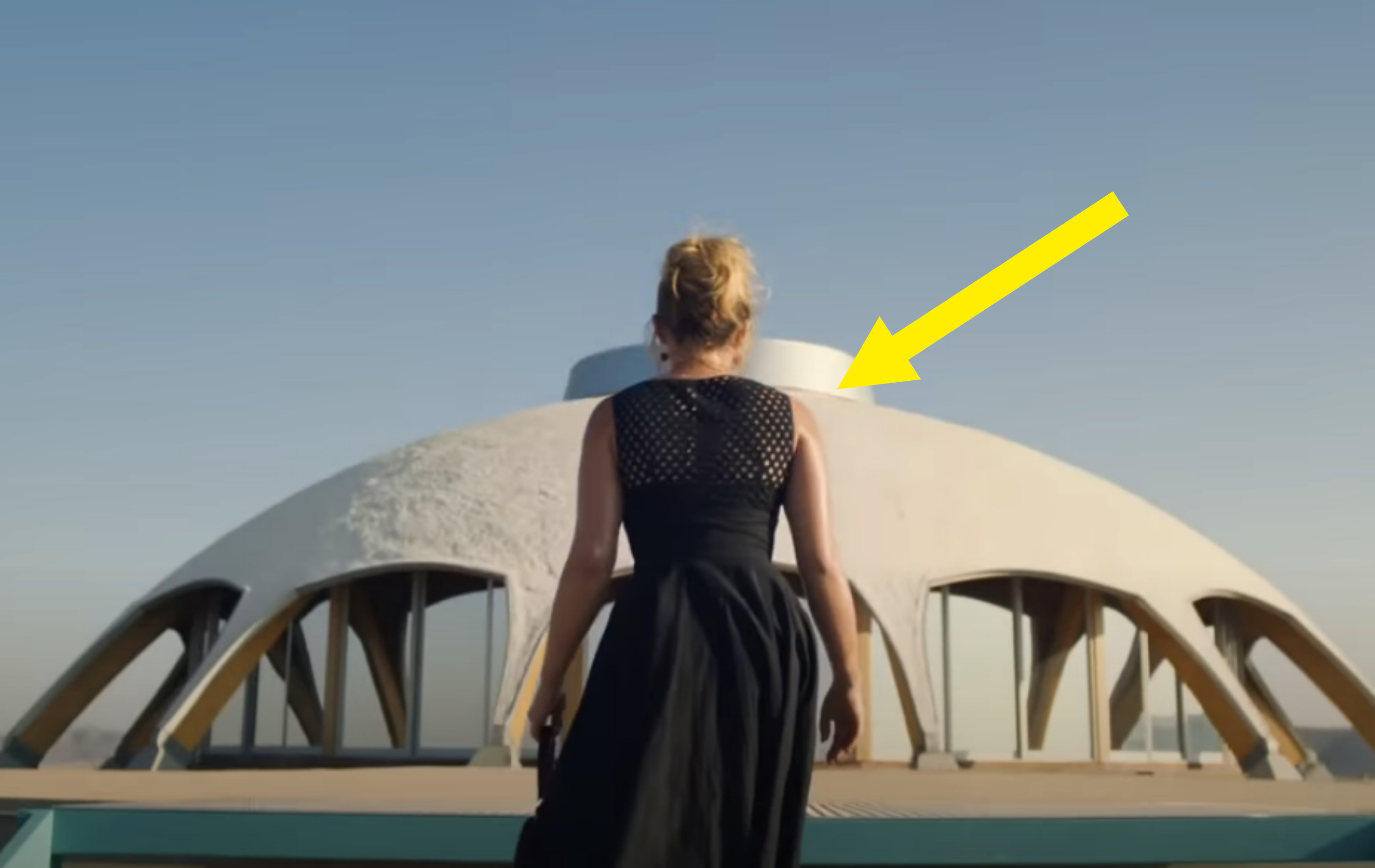 Alice wearing a simple, dark-colored dress and she walks toward a domed structure
