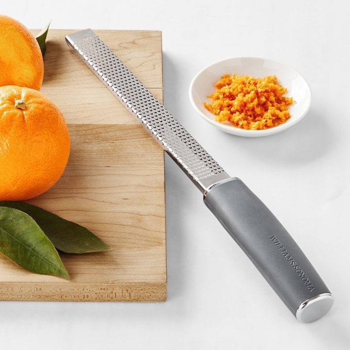 A microplane leaning against a wooden cutting board with oranges sitting on top of it. There is a bowl of zest.