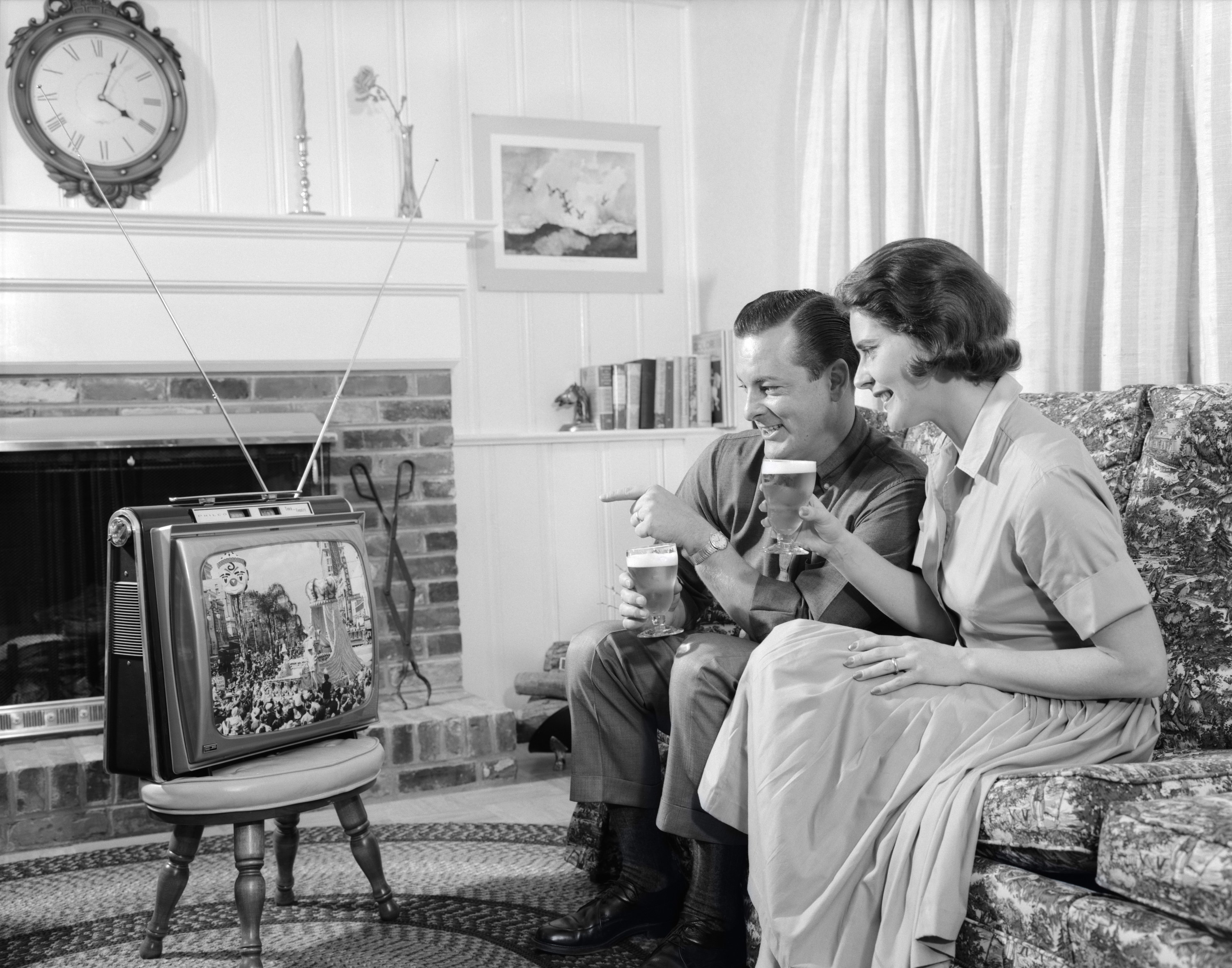 Black-and-white photo of a man and a woman sitting on a couch and looking at a small black-and-white TV on a stool