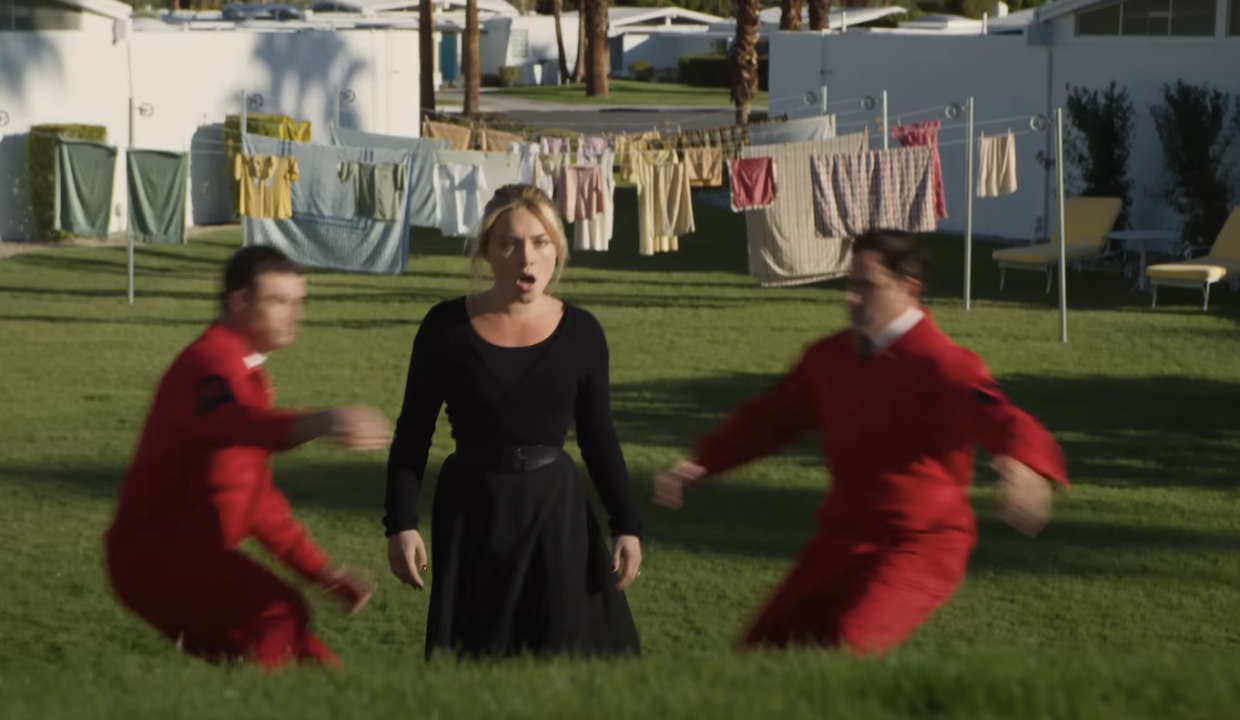 Two men in red jumpsuits reach out to grab Alice as she walks through a field full of laundry hung out to dry