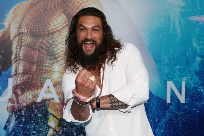 Jason Momoa smiling in front of an Aquaman poster