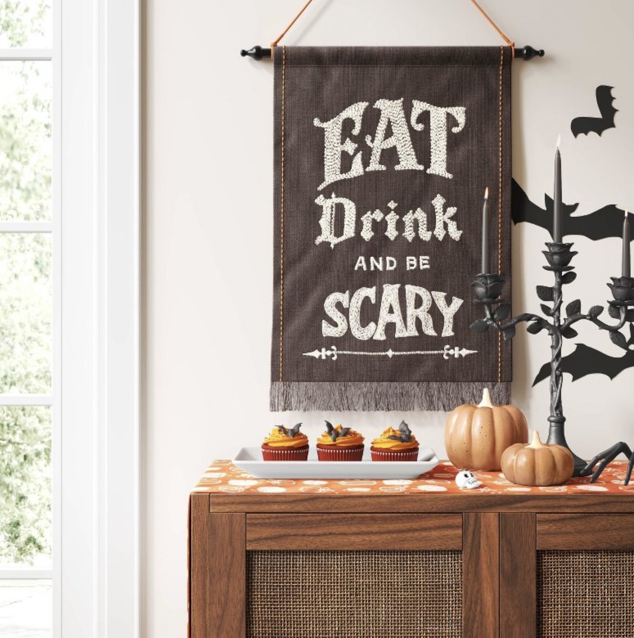 The small and medium ceramic pumpkins siting atop table next to platter of orange-frosted cupcakes. Banner on wall reads &quot;eat drink and be scary&quot;