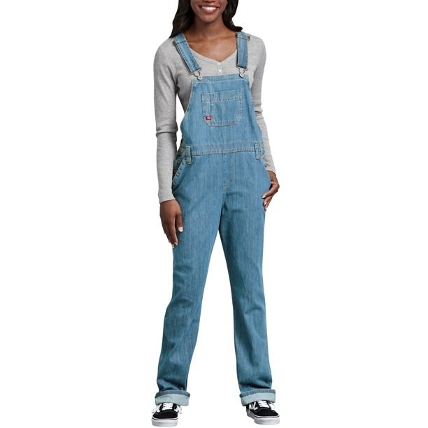 Model wearing light wash denim overalls with gray long-sleeved shirt