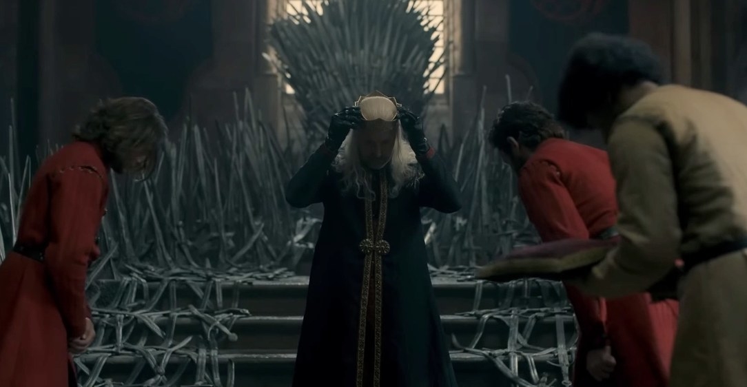Viserys stands in front of the throne and puts on his crown