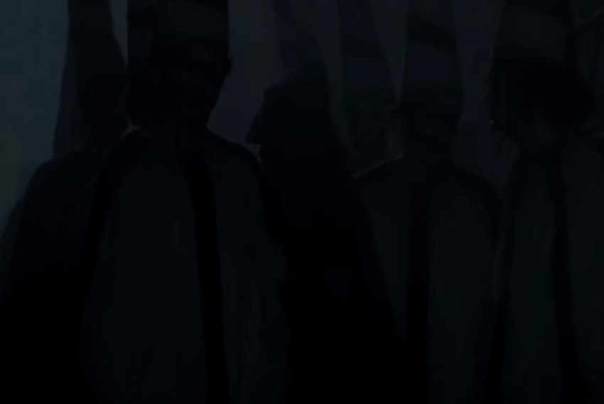 A hooded figure walks through men in tall hats