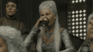A female character takes a sip of a drink and smirks