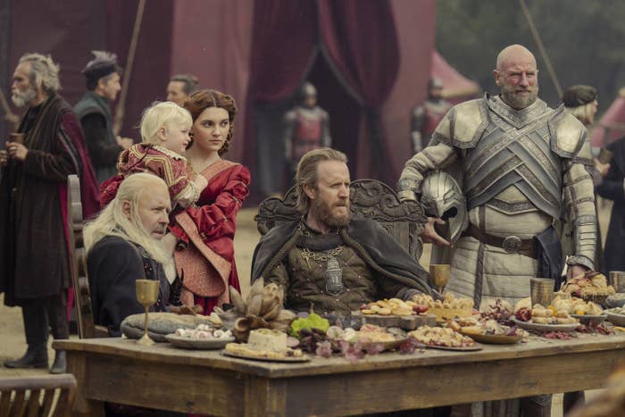 Some of the cast sit at an outdoor banquet table with lots of food