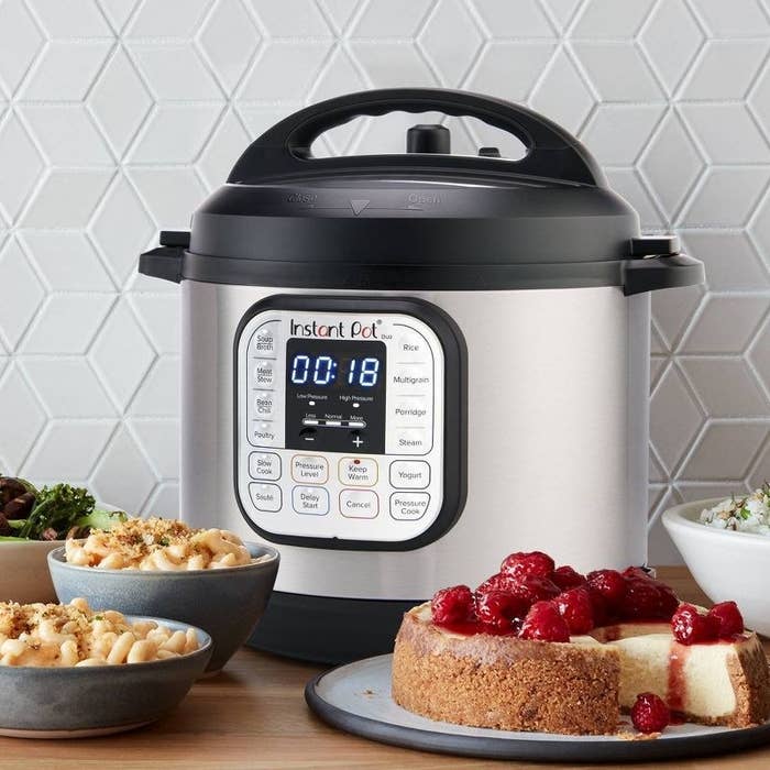 the silver pressure cooker on a counter surrounded by pasta, cheesecake and vegetables