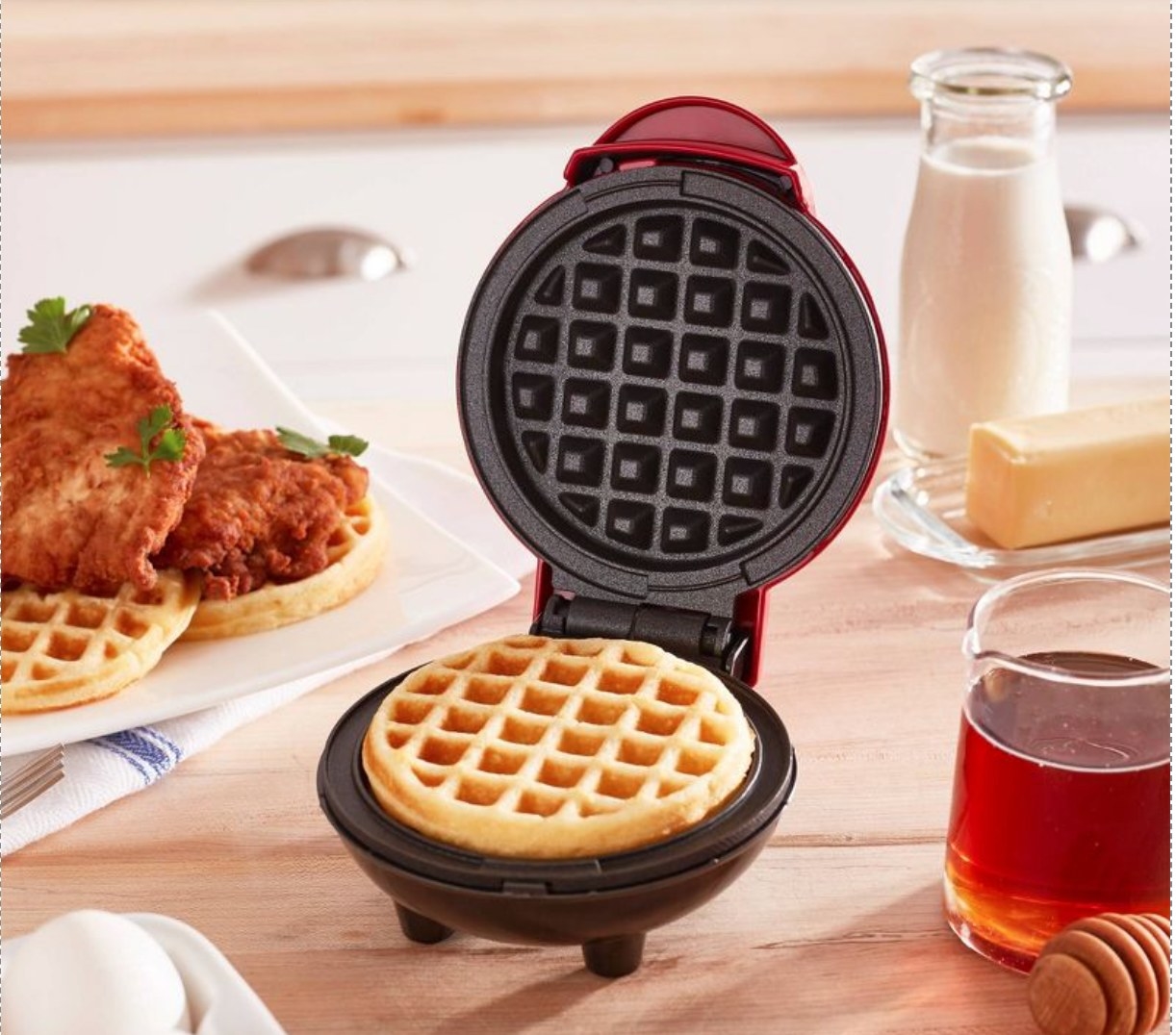 the small red waffle maker open on a counter to show the waffle inside. surrounding it are waffle ingredients and a plate of chicken and waffles