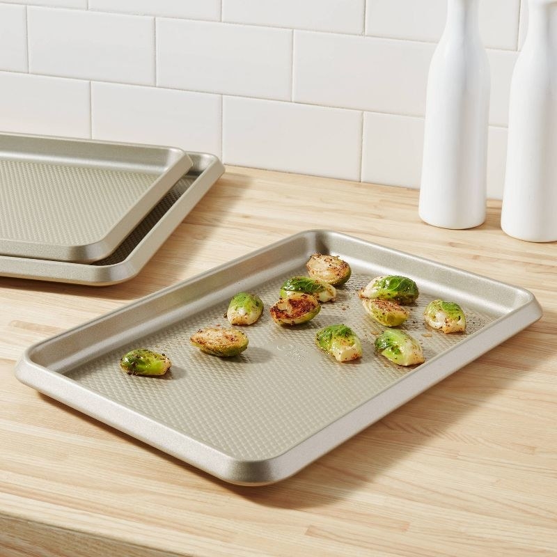 the gold baking sheet on a counter with brussels sprouts, and the other two sheets in the corner