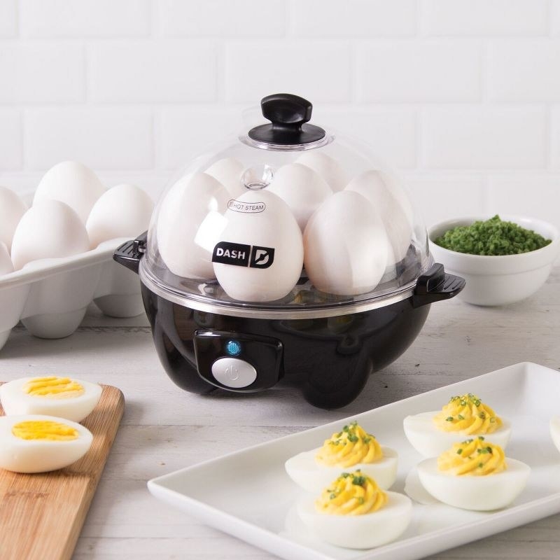 the black egg cooker filled with eggs, on a counter with more eggs and a plate of deviled eggs