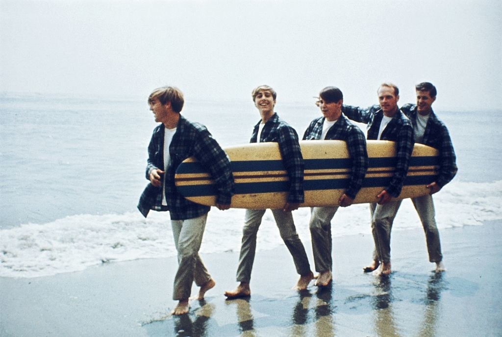 Group of guys holding a surf board and wearing suits on the beach