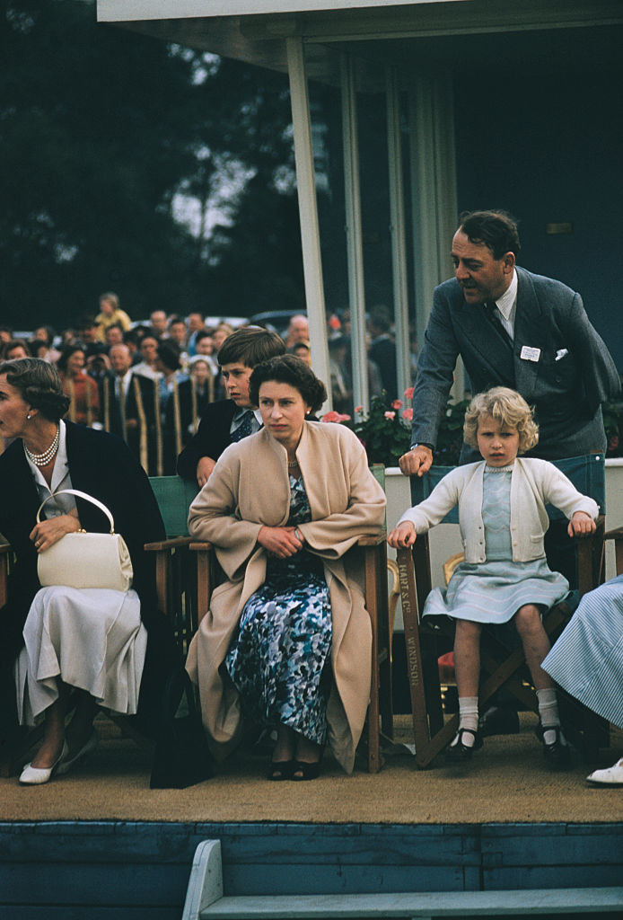 Elizabeth sitting at an outdoor event with her daughter, Princess Anne next to her, and Prince Charles behind her