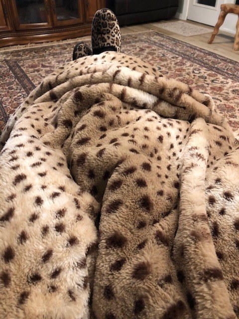 a reviewer photo of the cheetah print blanket over a person&#x27;s legs with cheetah slippers