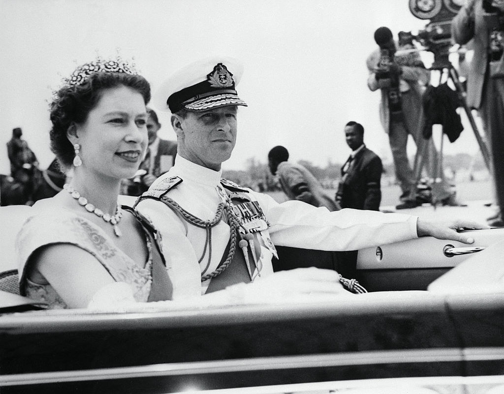 Queen Elizabeth, wearing a tiara, rides in a convertible with Prince Philip