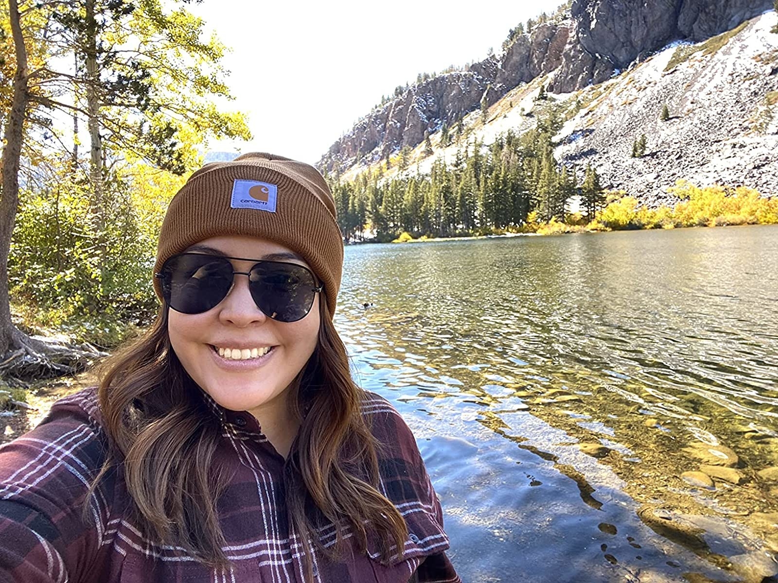 a reviewer photo of a person wearing the brown beanie and sunglasses, in front of a river and a mountain