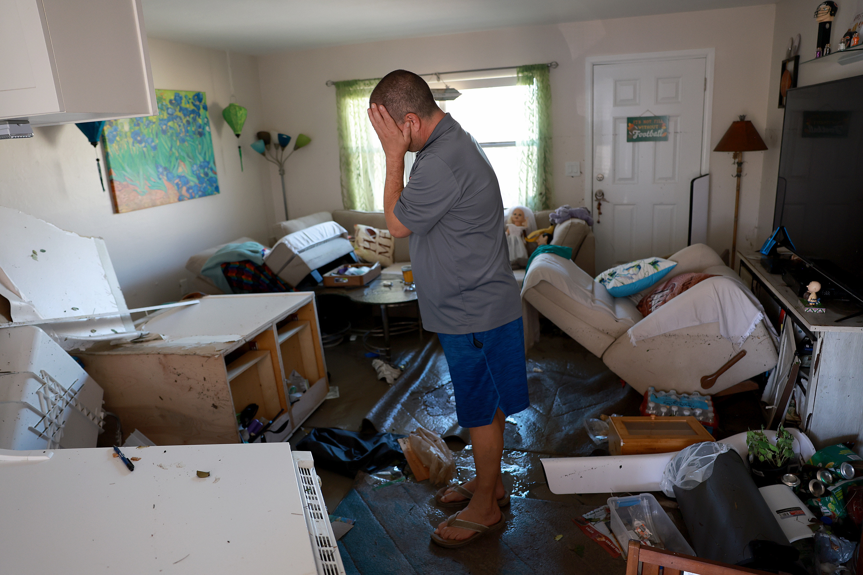A man wearing a gray shirt, blue shorts, flip flops stands in a cluttered, destroyed living room and holds his head in his hands and cries