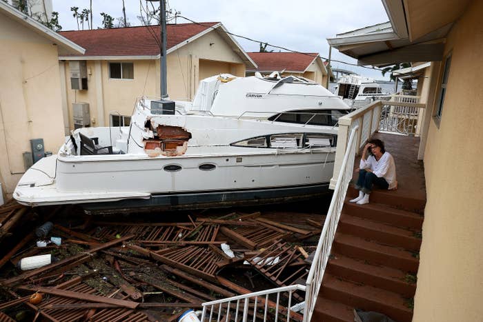 A large boat sits between an apartment complex over debris of a broken fence while a person sits on top of an outdoor staircase, holding their head in their hand