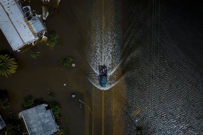 An aerial view of a truck driving down a flooded area, causing waves as it progresses with mobile homes, sunlight reflects off the ripples in the water