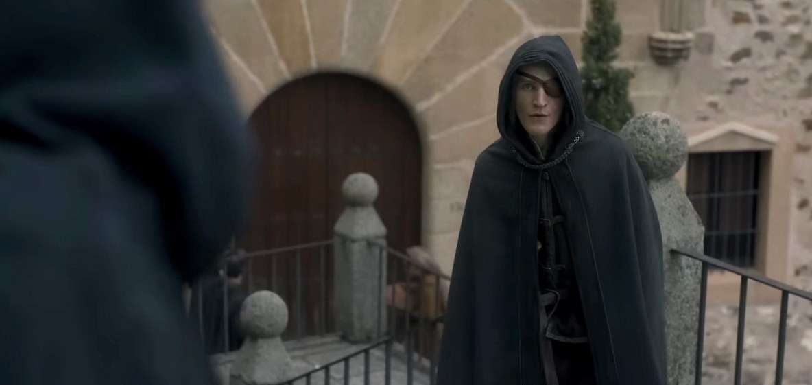 Aemond wears a hood and stands facing another hooded figure