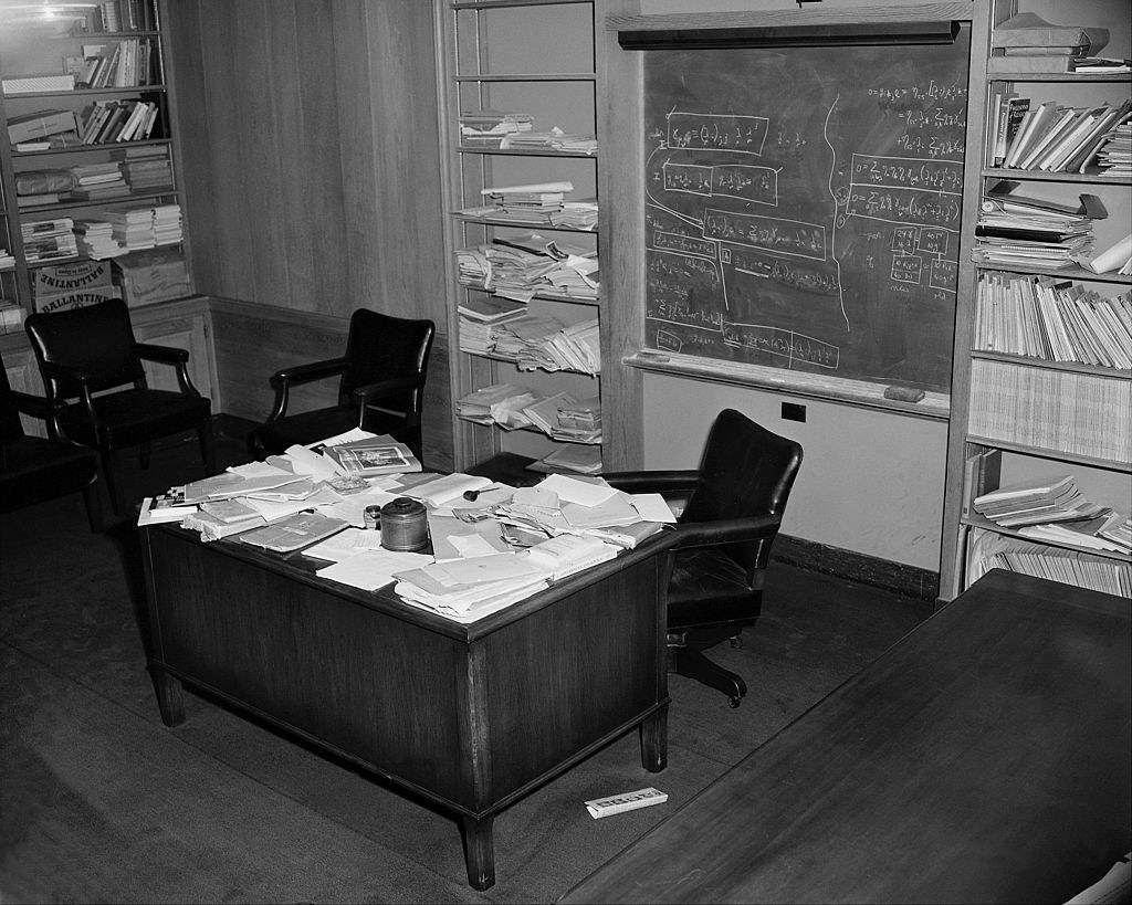 Black-and-white photo of a messy desk covered in papers and journals, with a blackboard behind it with lots of writing, and shelves of journals, notepads, and books on either side