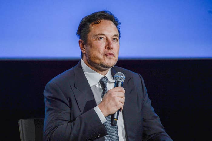 Elon siting onstage in a suit and tie as he holds a mic
