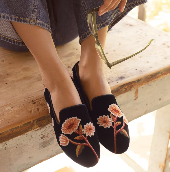 a model wearing black slip-on flats with pink embroidered flowers on them