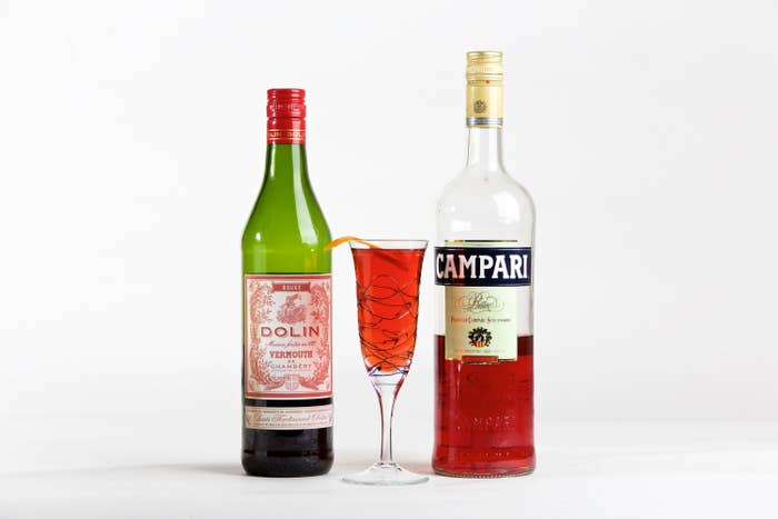A bottle of Dolin sweet vermouth next to a flute glass with a negroni sbagliato in it and an orange peel, next to a bottle of Campari
