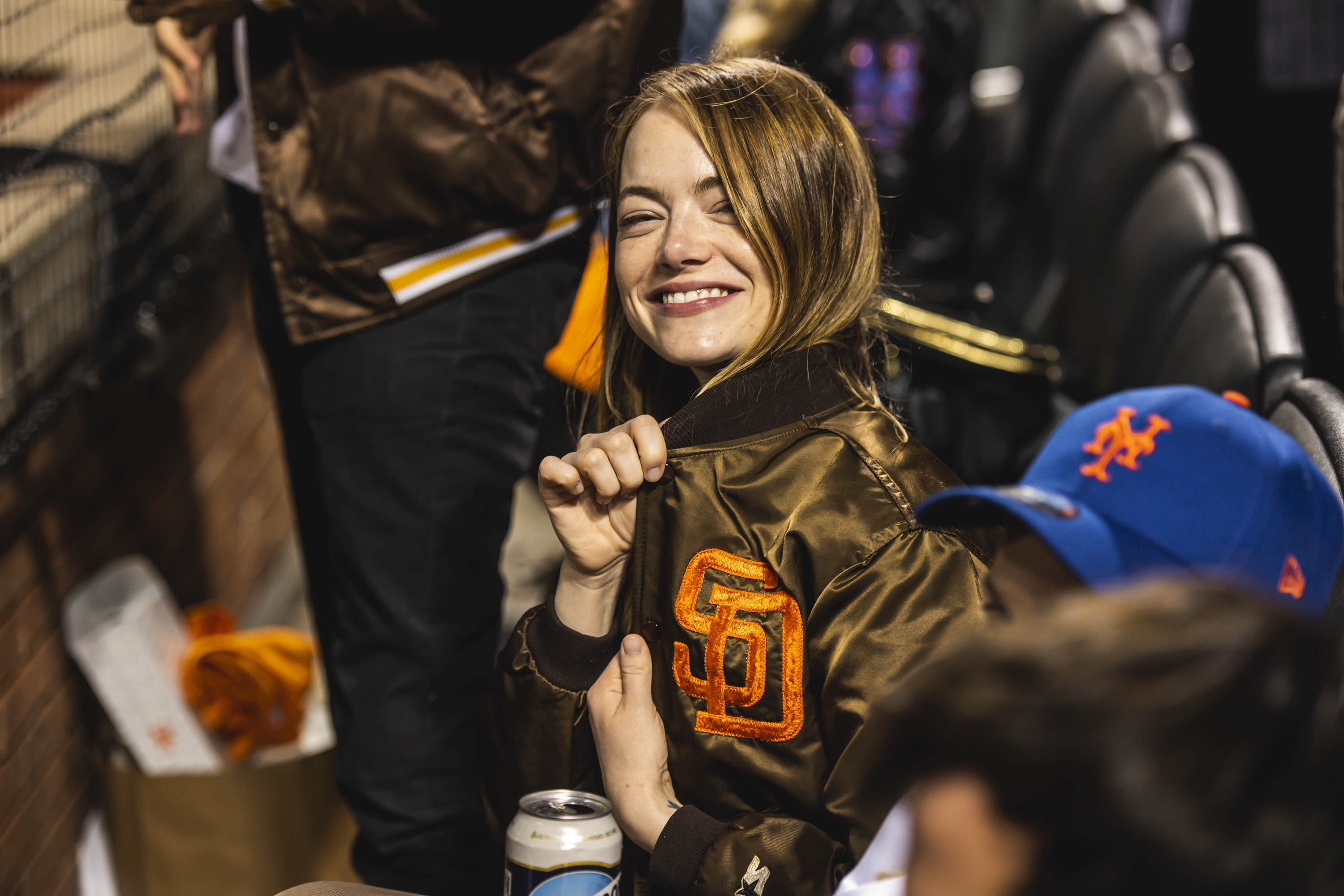 Emma sits in the crowd and shows off the baseball logo on her jacket