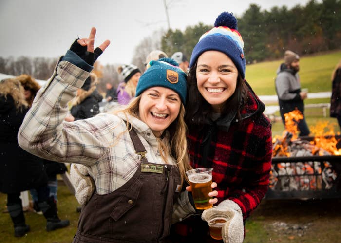 Two smiling women in flannel wearing beanies near a fireplace during the daytime