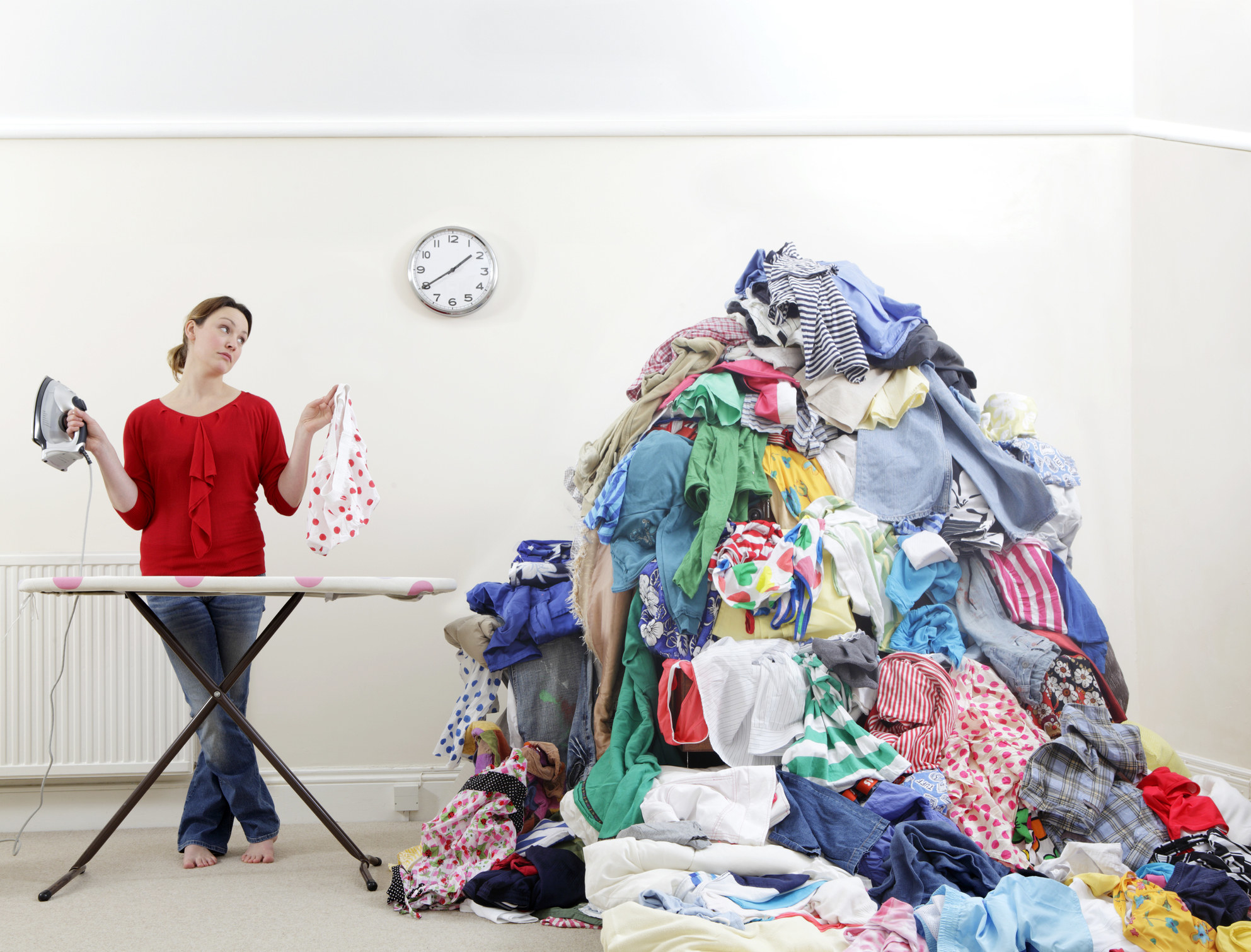 A woman standing at an ironic board and holding up an iron and a piece of clothing while looking at a mountain of clothing next to her