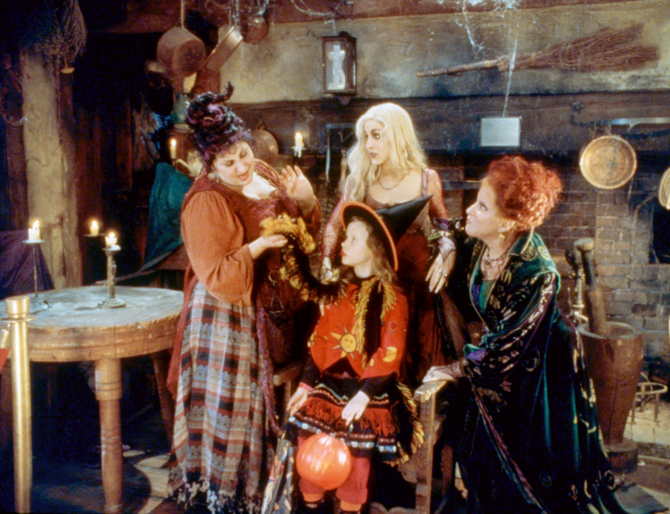 The Sanderson sisters standing around a young girl carrying a pumpkin candy holder
