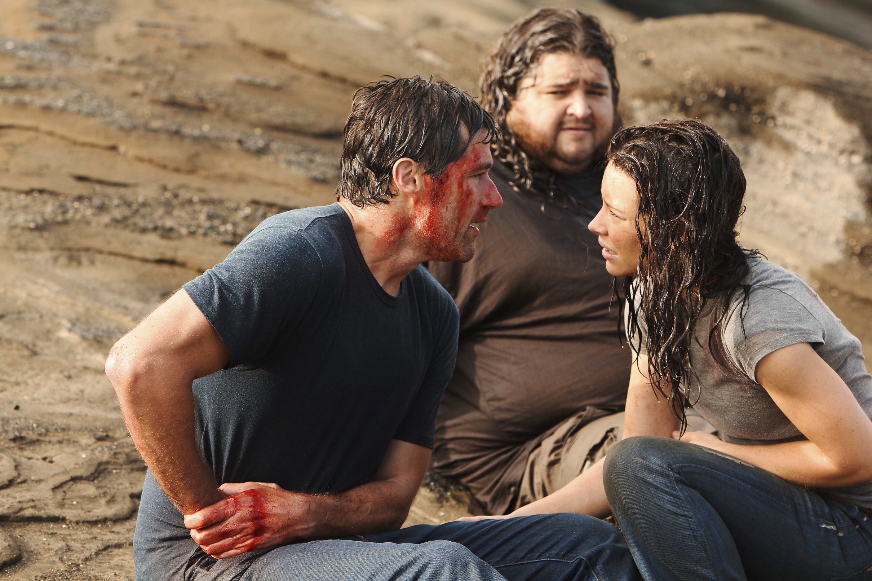 three of the characters sitting on a beach, exhausted, one with blood all over his face