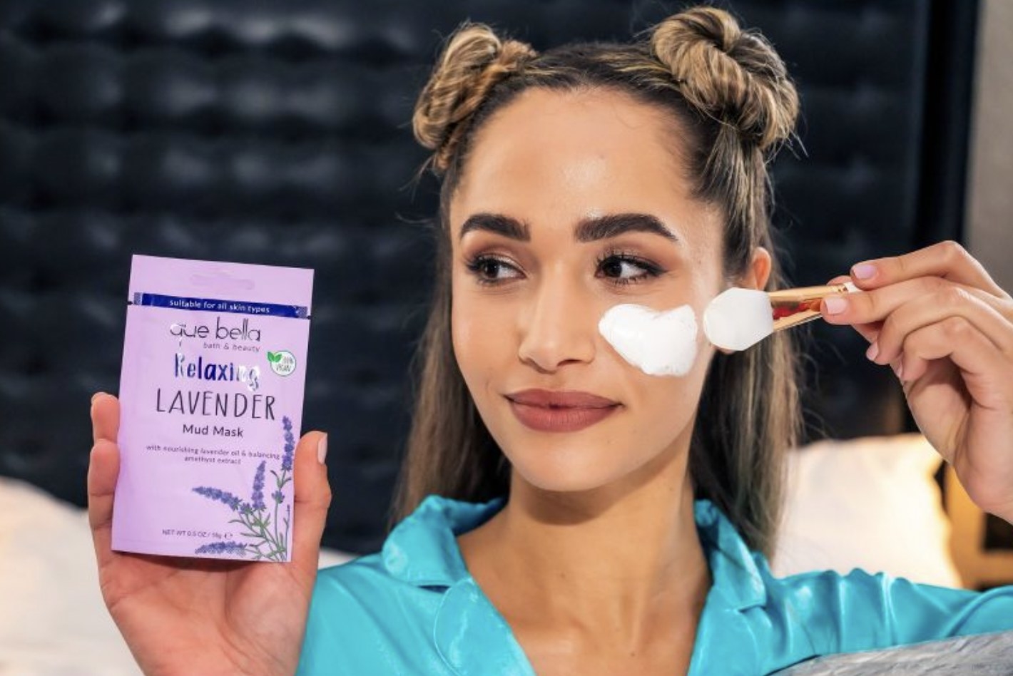 Model applying the mud mask to face and holding up small lavender mud mask package