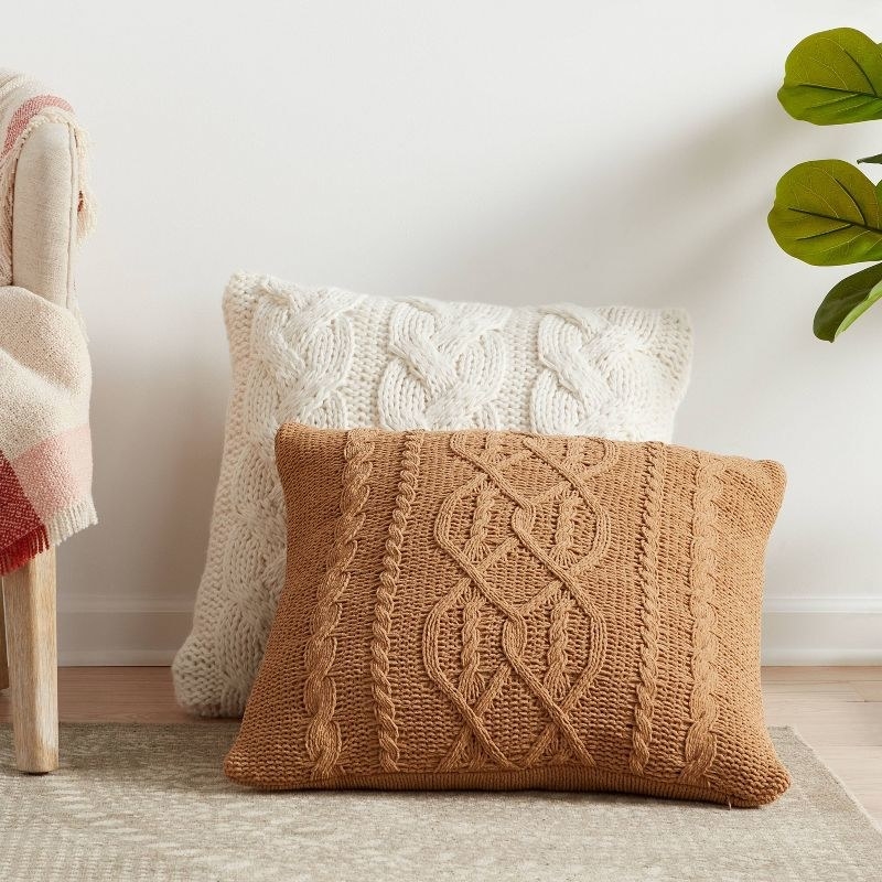 the tan and white cable knit pillows on the floor next to a chair and a plant