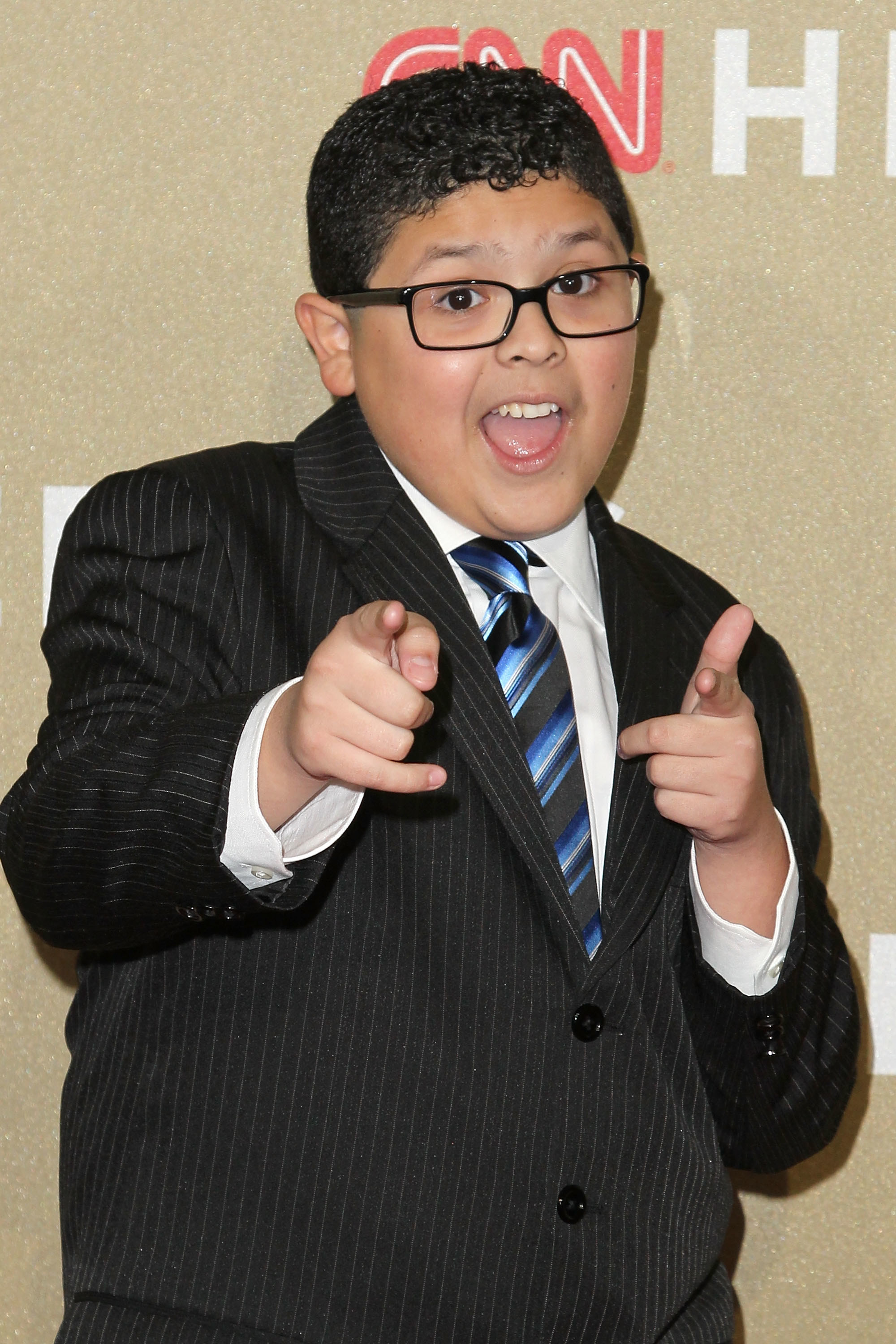 Rico wearing eyeglasses and a suit pointing finger guns at the camera