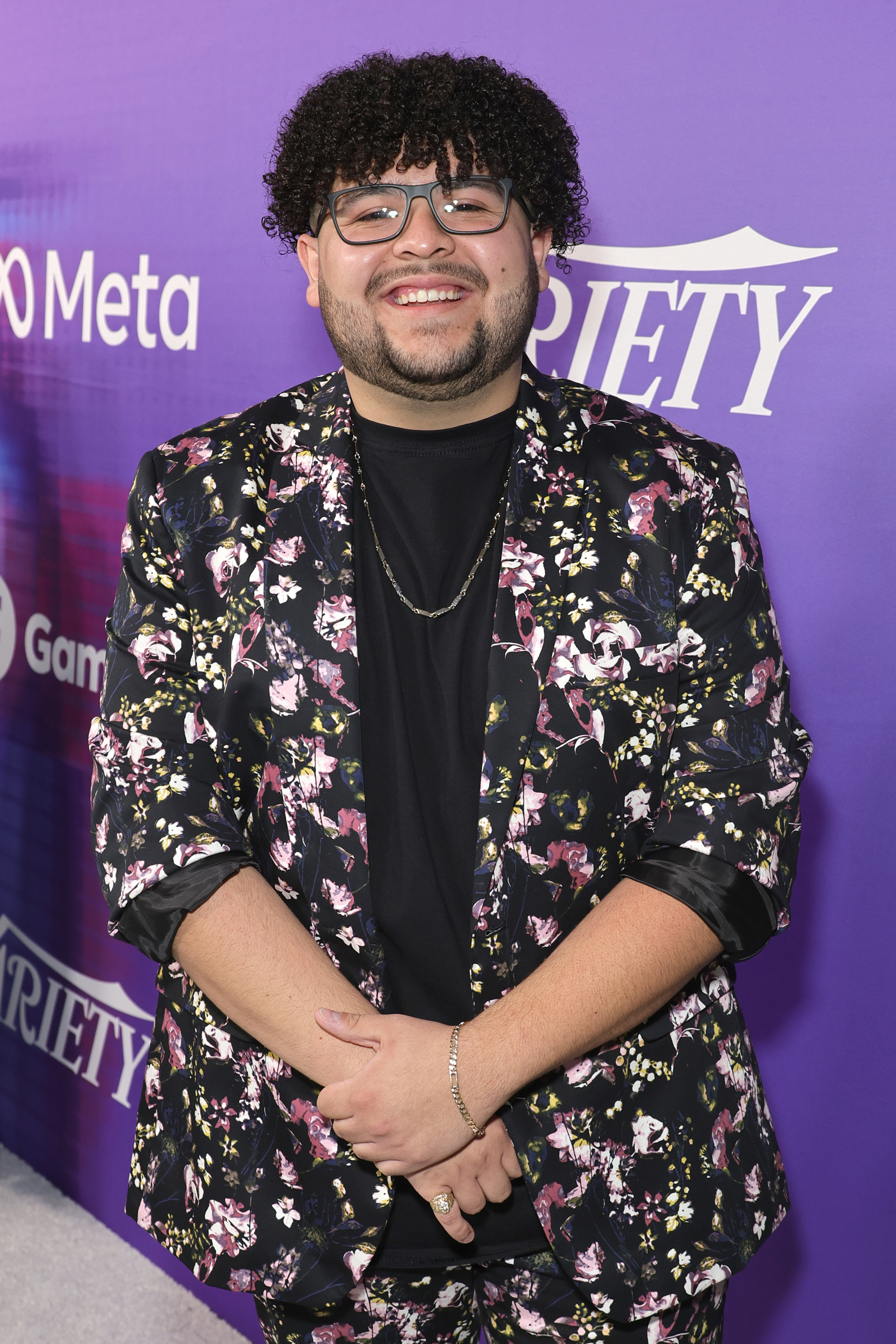 Rico in a floral suit, eyeglasses, and long curly hair smiling at an event