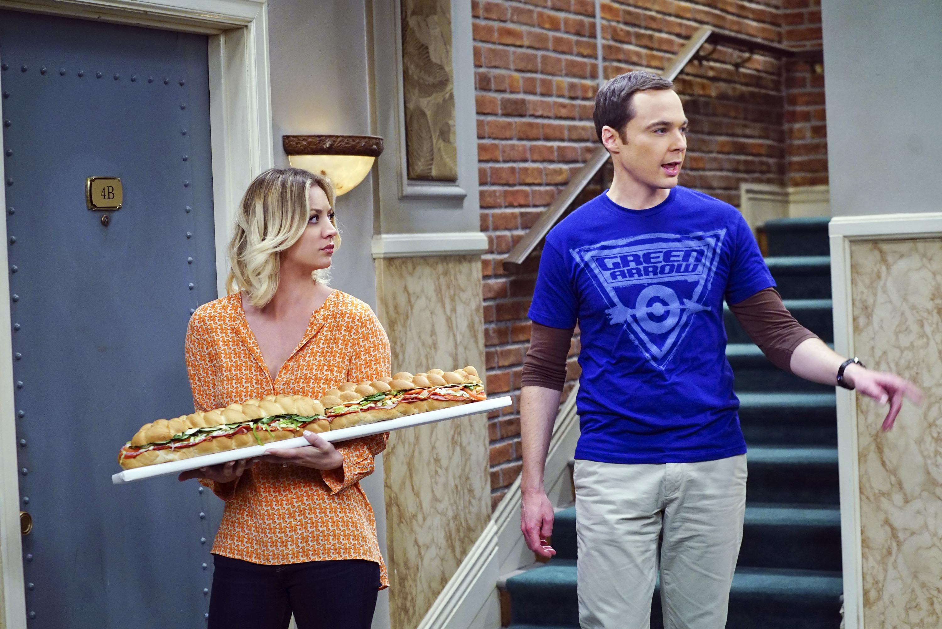 Kaley holding a long sub and standing next to Jim Parsons in the apartment building hallway in a scene from The Big Bang Theory