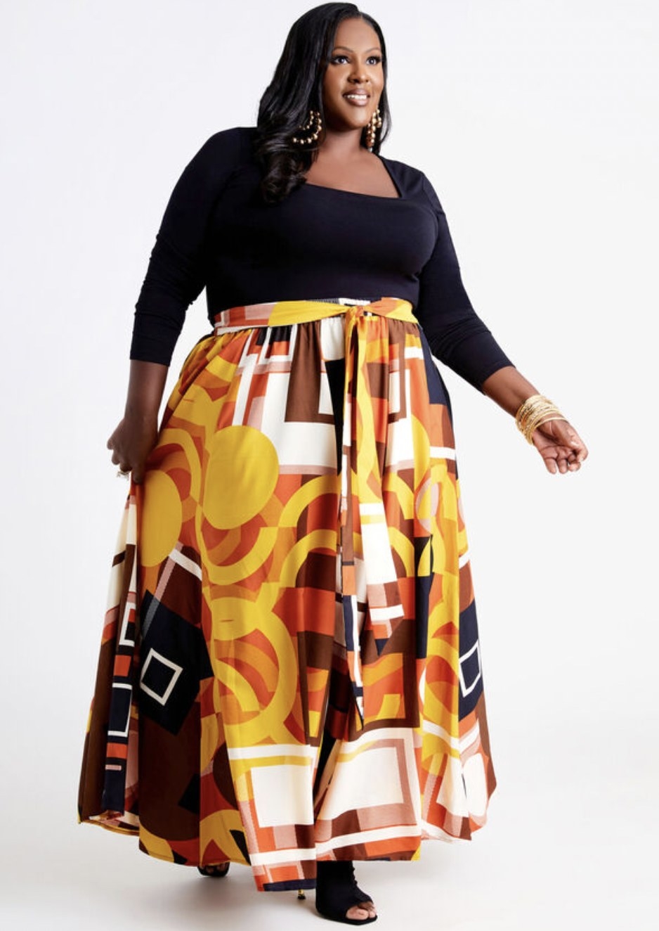 Model wearing the geometric orange and red toned print skirt with belt tied at waist, paired with black long sleeve tee