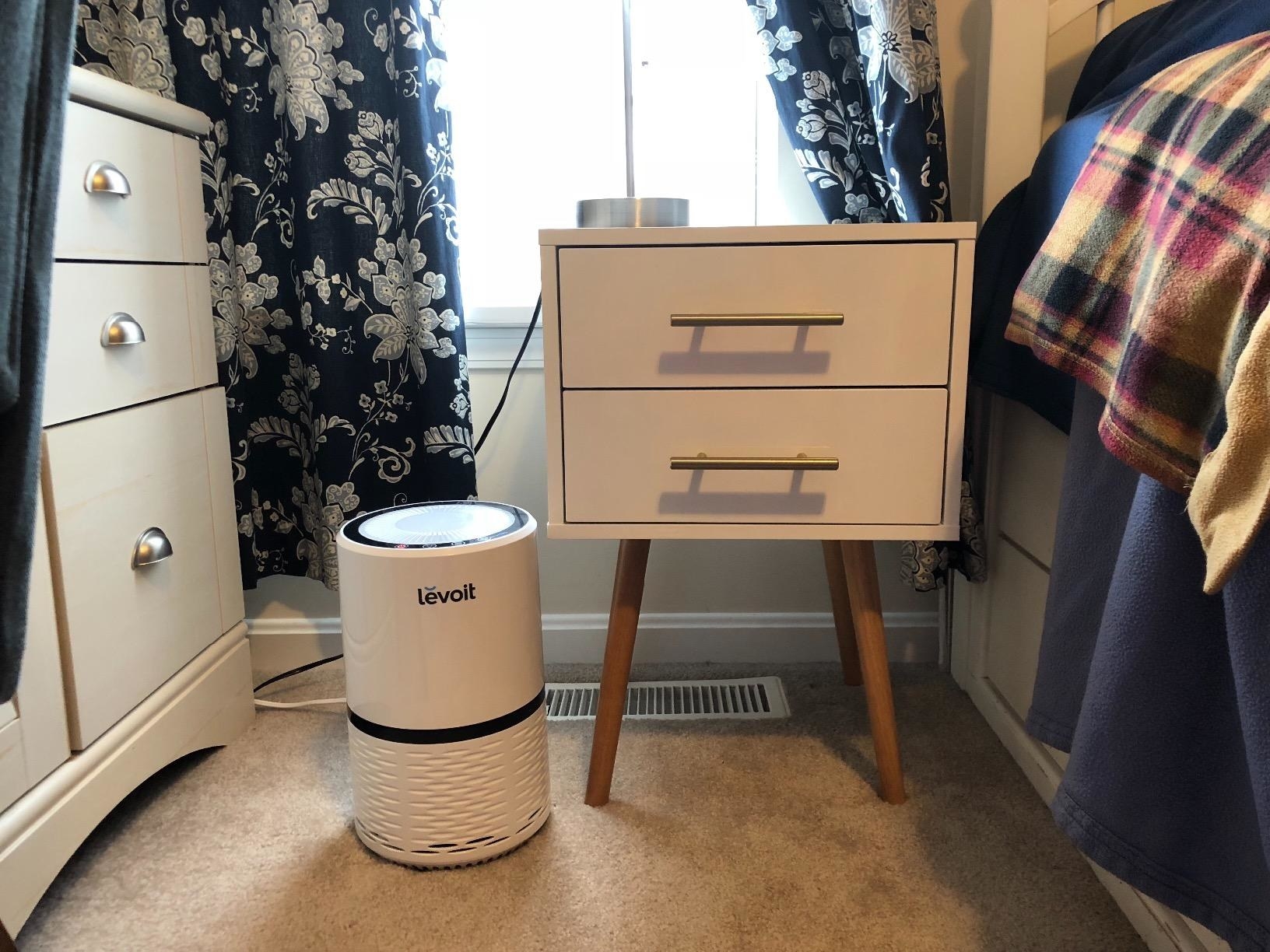 A reviewer&#x27;s white air purifier in their bedroom, next to a nightstand to show its small size