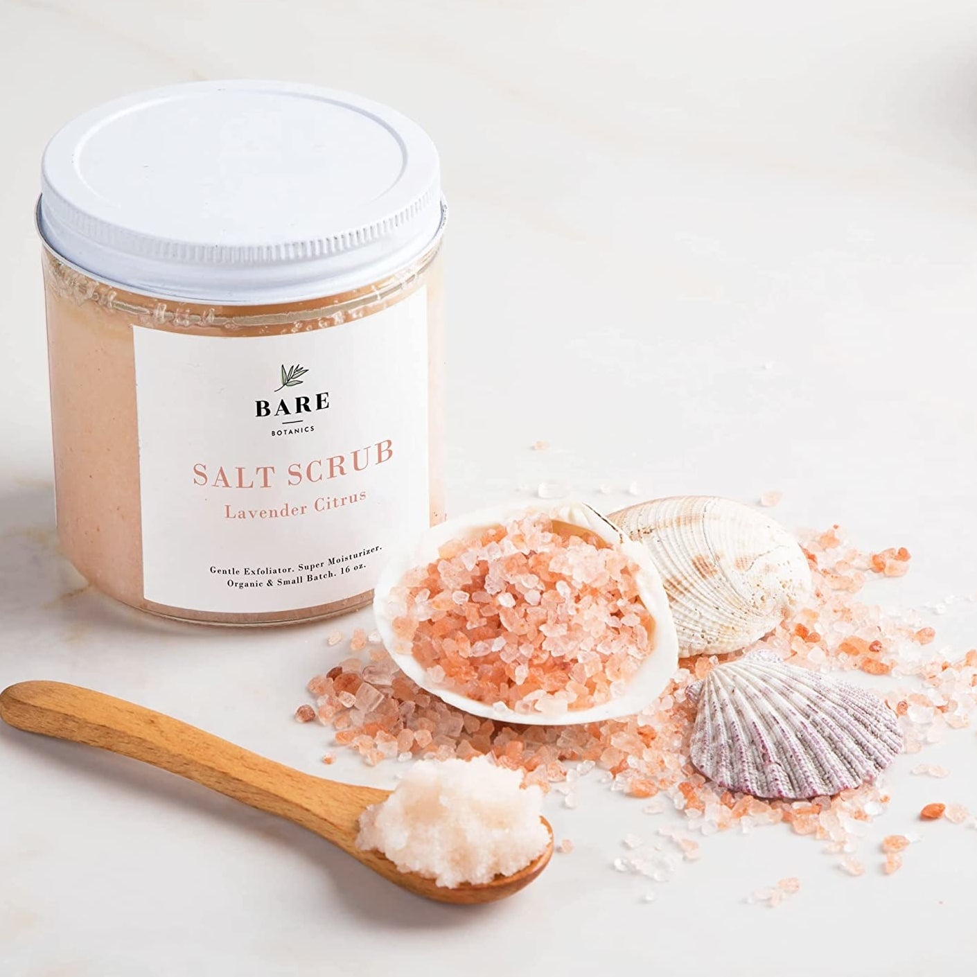 the tub of salt scrub next to a seashell full of pink salt and a spoon of the scrub