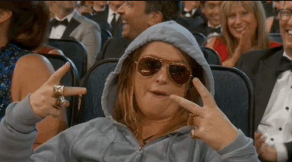 A person sitting in an audience wearing a hoodie and sunglasses and giving the V sign